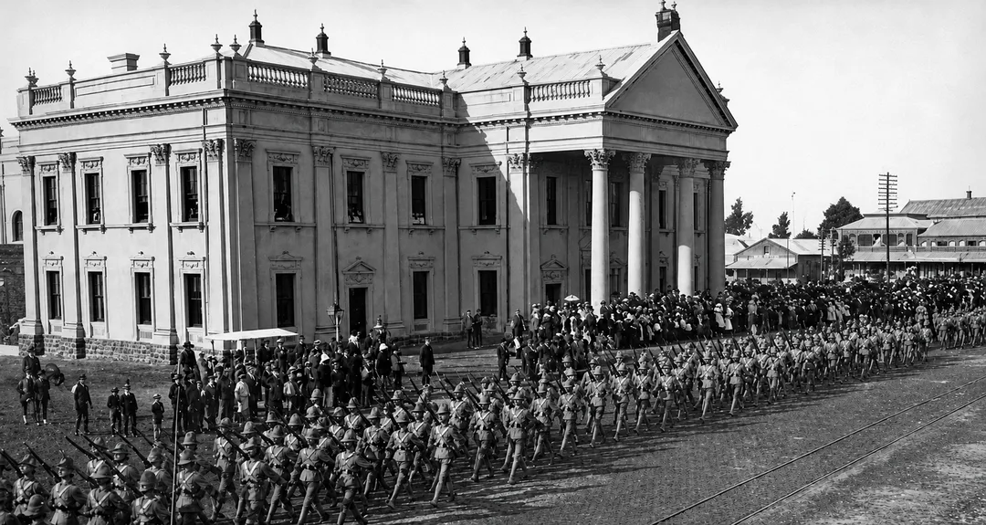 Historic black-and-white photograph of Kimberley Town Hall in September 1899, with a large formation of uniformed soldiers marching across the foreground and crowds gathered outside the newly completed Victorian building. The Post Office and other early Kimberley buildings are visible in the background.