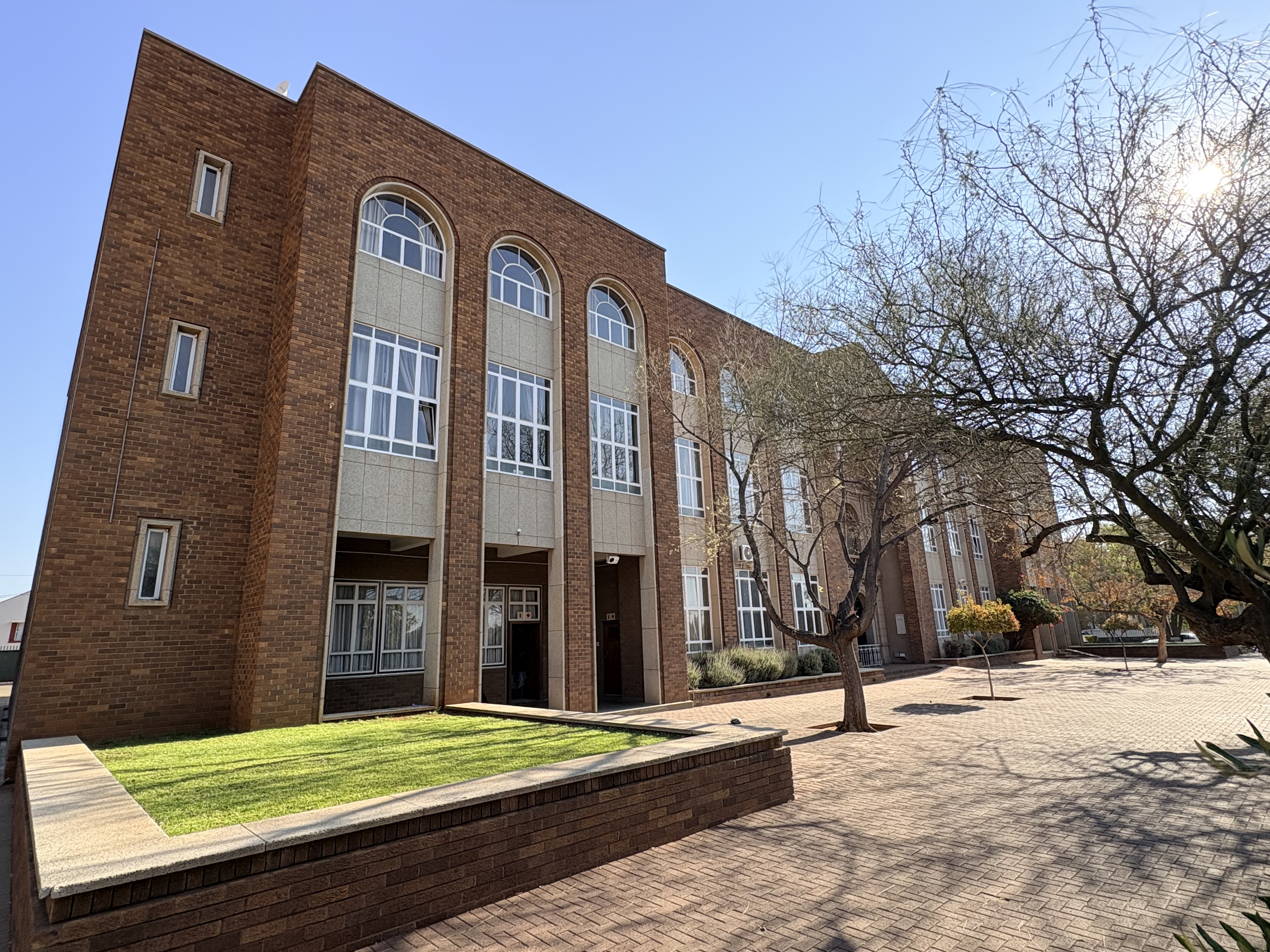 Oblique front view of St Patrick’s CBC Kimberley showing a long red-brick school building with tall arched windows, a paved courtyard, lawns and mature trees in the foreground.