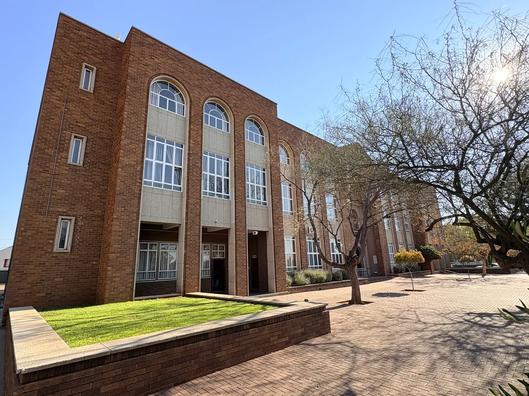 Oblique front view of St Patrick’s CBC Kimberley showing a long red-brick school building with tall arched windows, a paved courtyard, lawns and mature trees in the foreground.