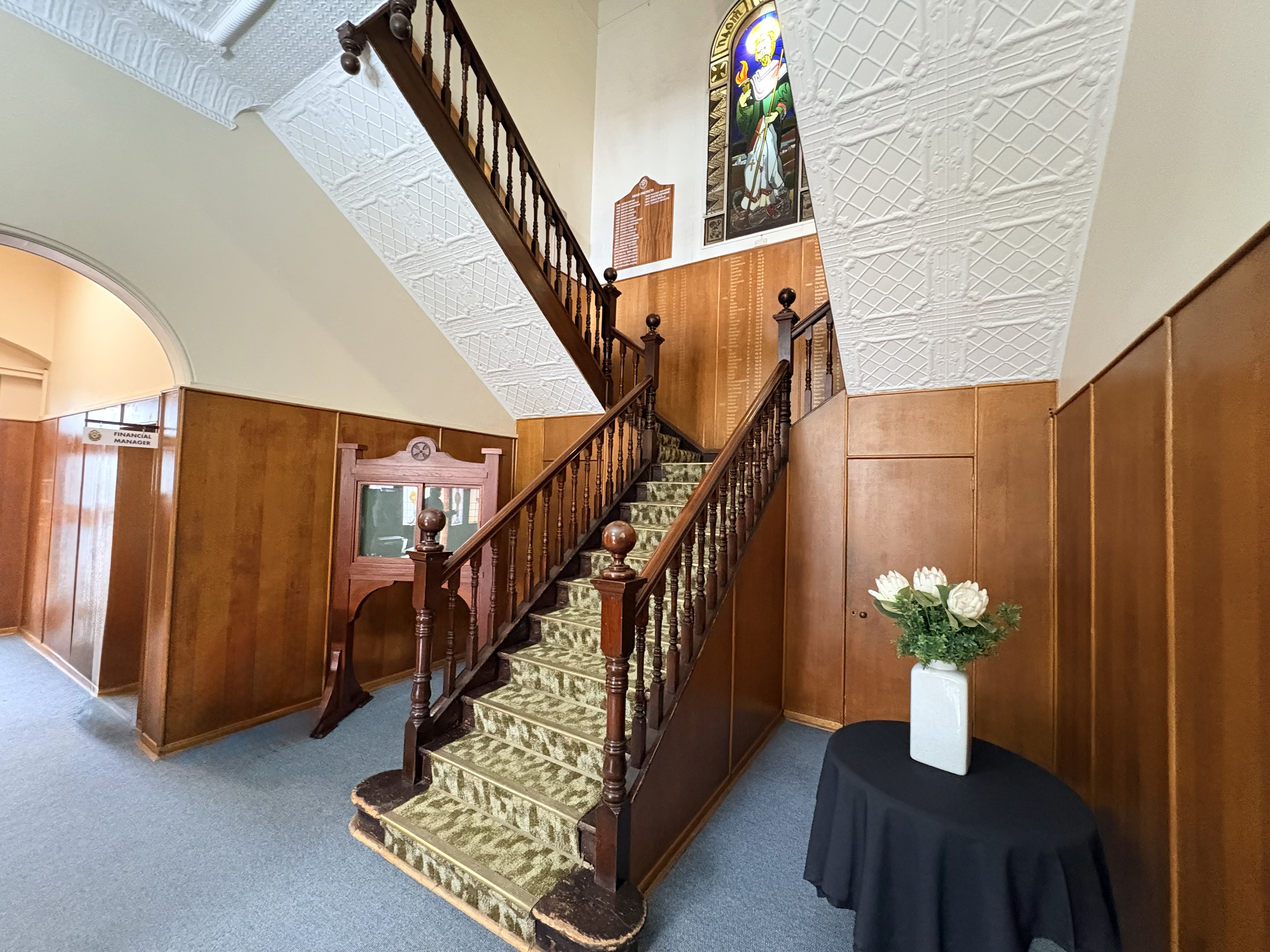 Entrance hall beneath the War Memorial Clock Tower at St Patrick’s CBC in Kimberley, showing a timber staircase, decorative ceiling, stained-glass window and memorial boards.