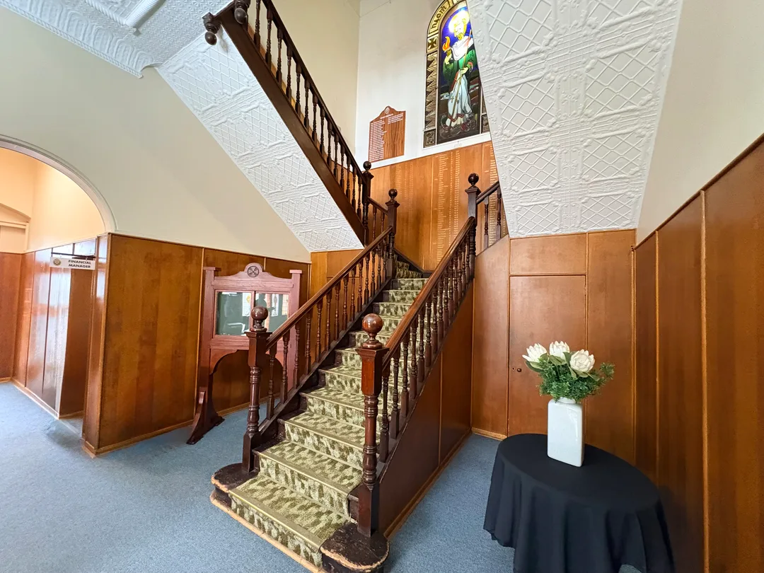 Entrance hall beneath the War Memorial Clock Tower at St Patrick’s CBC in Kimberley, showing a timber staircase, decorative ceiling, stained-glass window and memorial boards.
