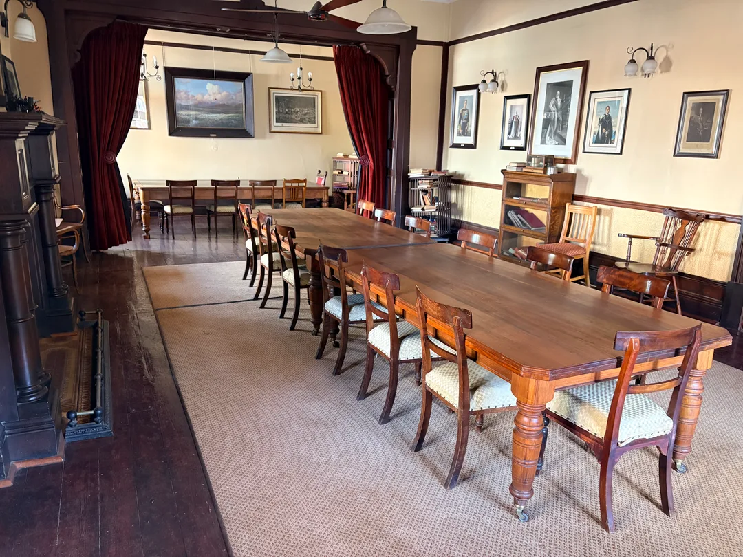 Reading Room on the upper floor of the Kimberley Club, with long timber tables, historic chairs, fireplace, framed photographs and deep red curtains.