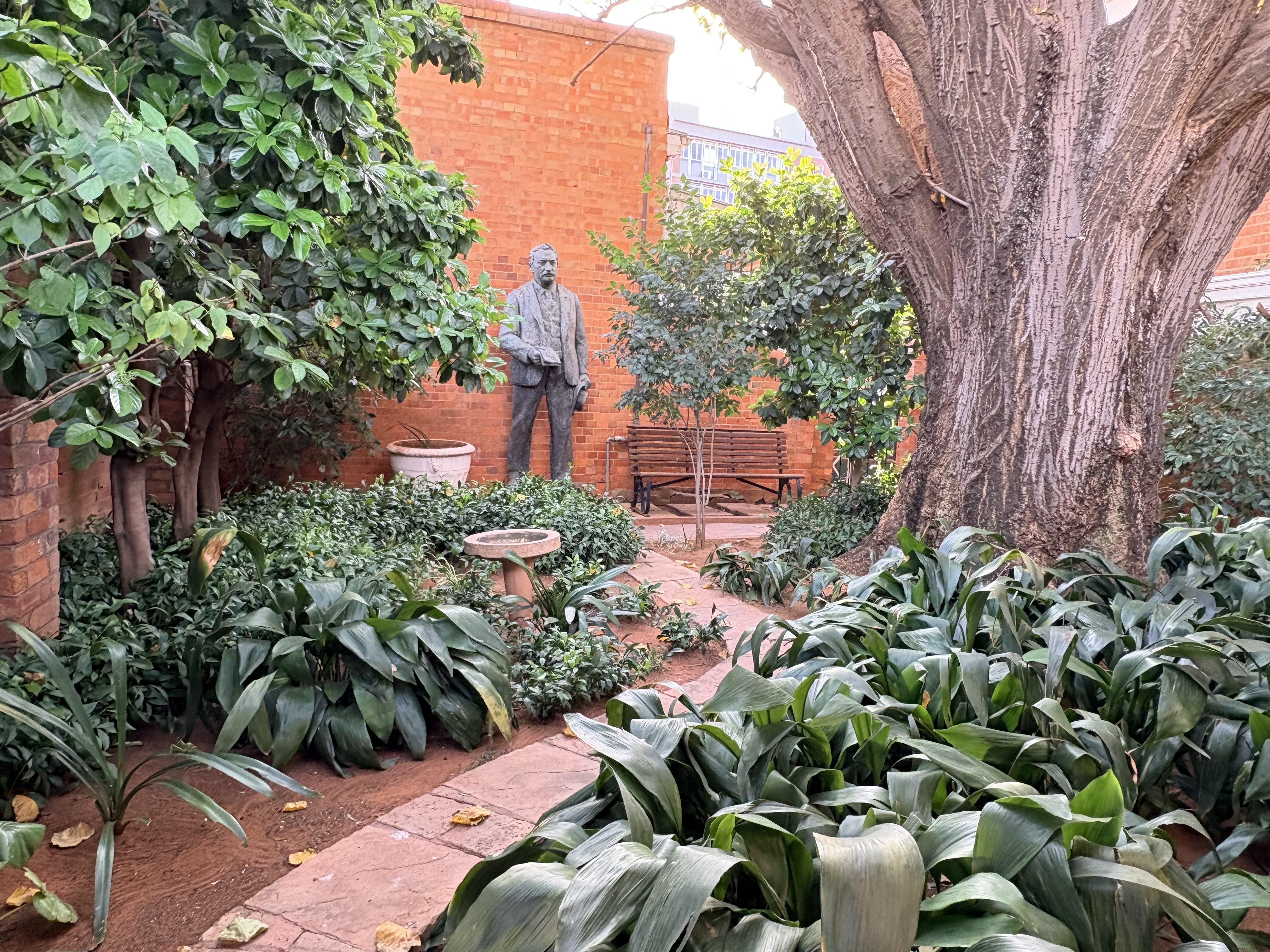 Garden courtyard at the Kimberley Club in Kimberley, with mature trees, planted beds, a bench and a historic statue reportedly relocated from Mahikeng Station.