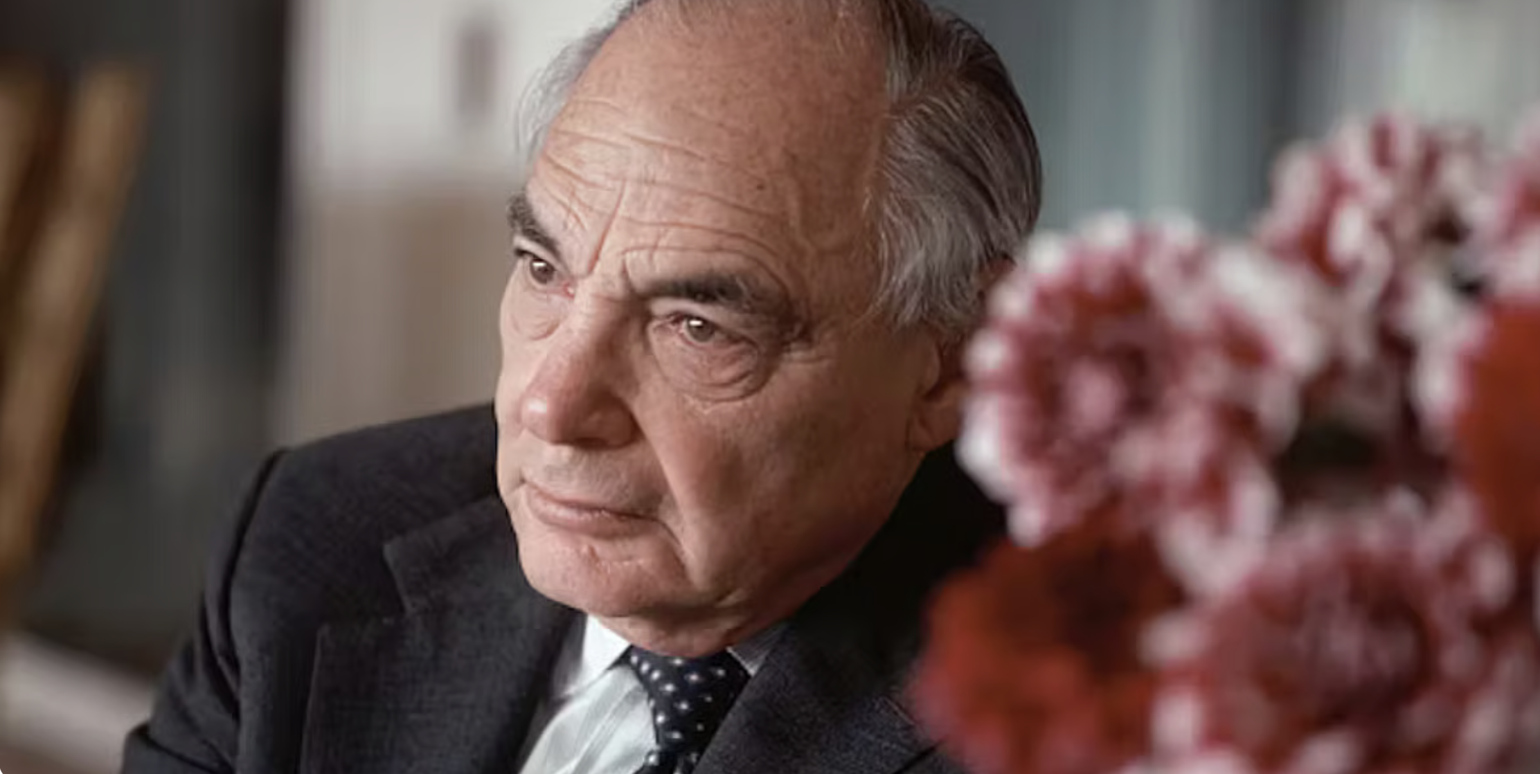 Colour portrait of Harry Oppenheimer in later life, wearing a dark suit and tie, photographed indoors with flowers in the foreground.